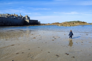 Un enfant se tient au premier plan, au second plan la mer, des rochers et les remparts de Saint-Malo